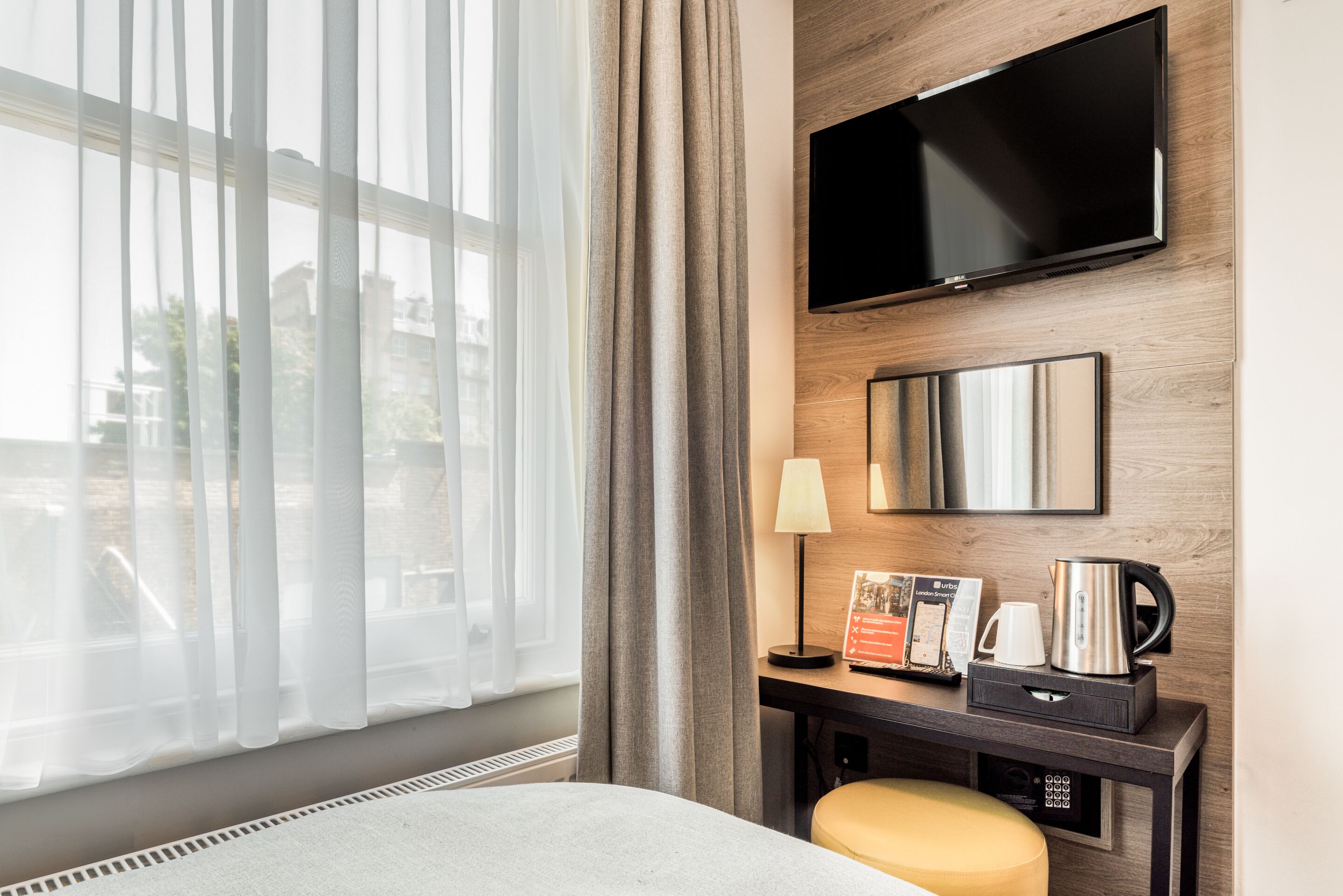 A hotel room with a bed, a desk with a lamp, a coffee maker, a television, and a window with curtains.