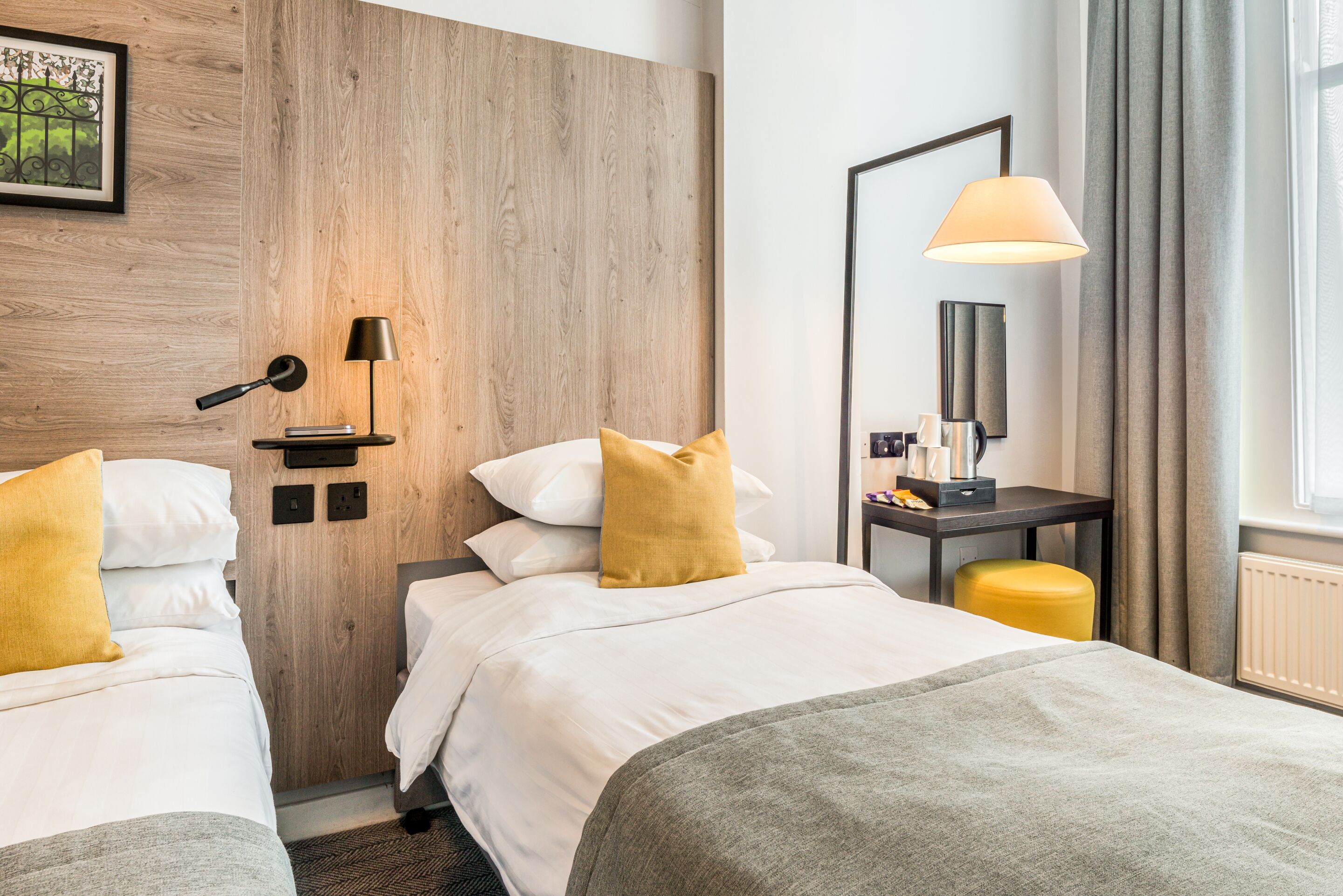 A hotel room with two beds, a wooden accent wall, a desk with a lamp, and a window with curtains.