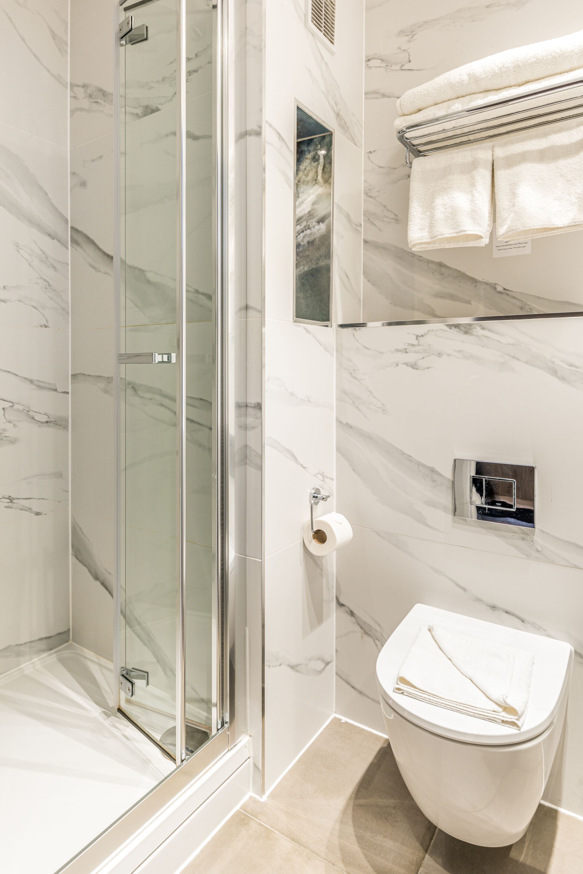 A modern bathroom with a glass-enclosed shower, a toilet with a towel on top, and marble walls.
