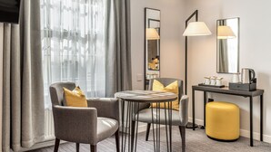 A modern hotel room with a grey sofa, a round table, a yellow ottoman, a desk with a lamp, and a large mirror. - The Princes Square Hotel (London)