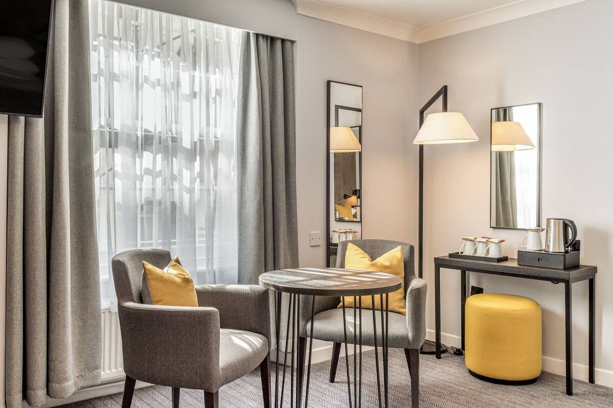 A modern hotel room with a grey sofa, a round table, a yellow ottoman, a desk with a lamp, and a large mirror.