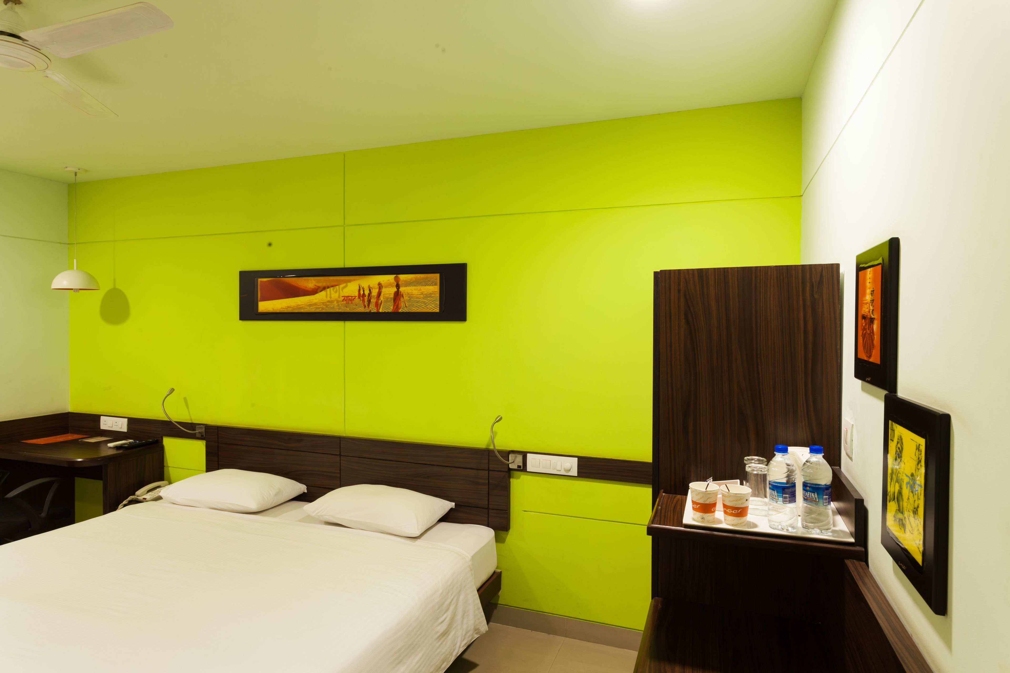 A hotel room with a bed, a desk, a chair, a nightstand, and a wall with artwork.