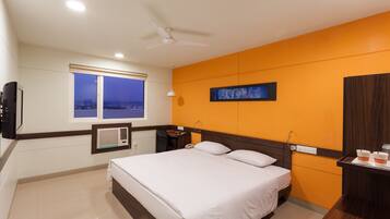 Habitación de hotel con cama, escritorio, silla, ventana, ventilador de techo y televisor de pared.