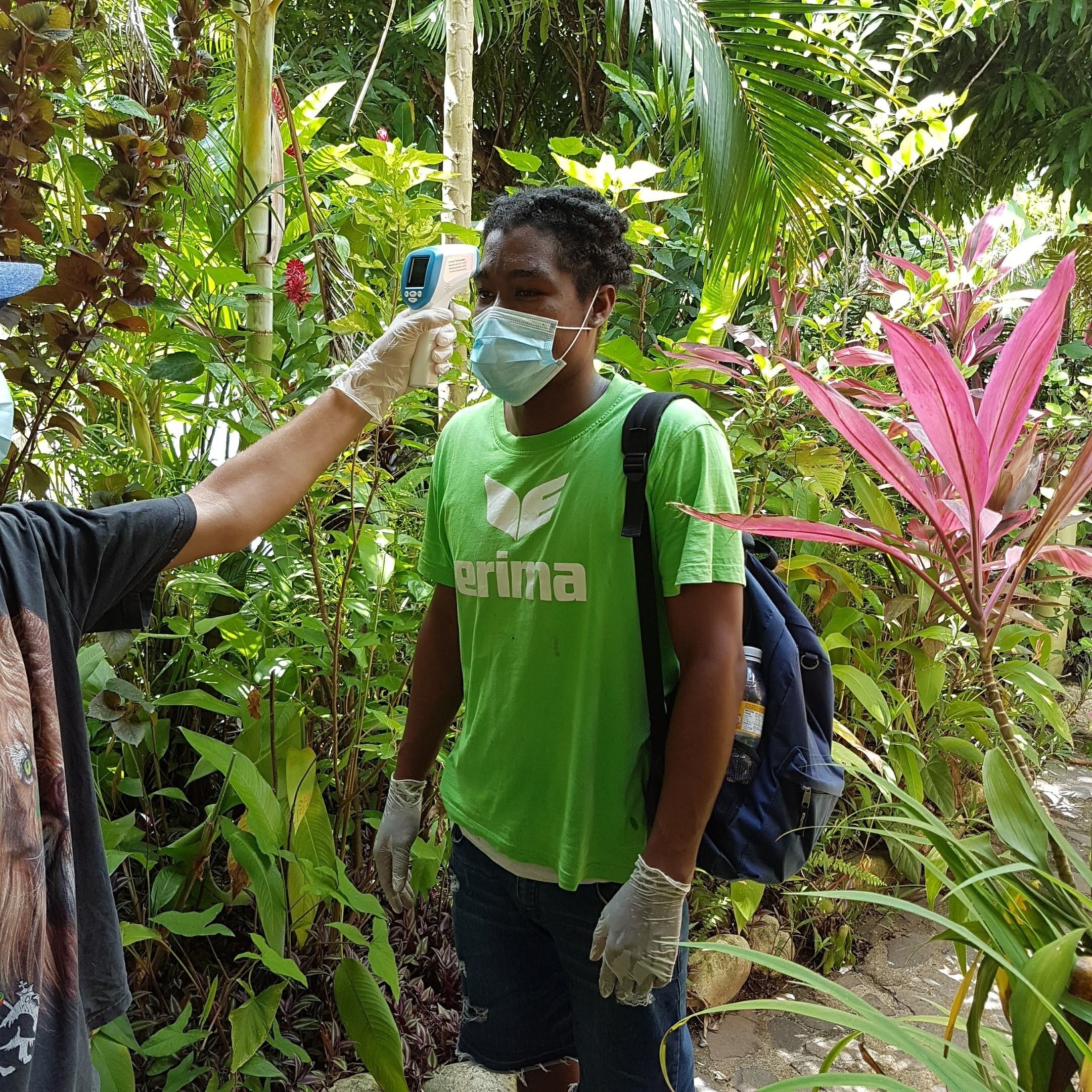 A person wearing a green shirt with the word "erima" and a backpack, standing in a lush, green outdoor area.