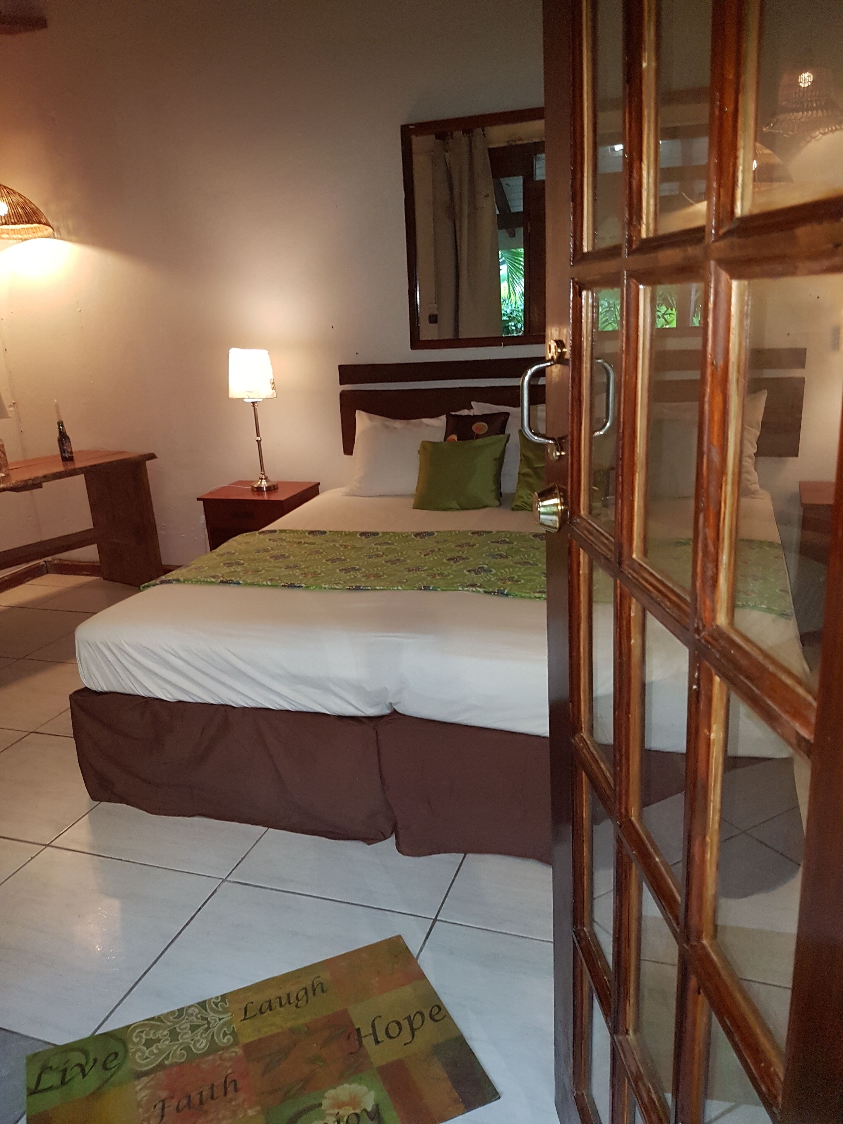 A hotel room with a bed, a nightstand, a lamp, a mirror, and a door.