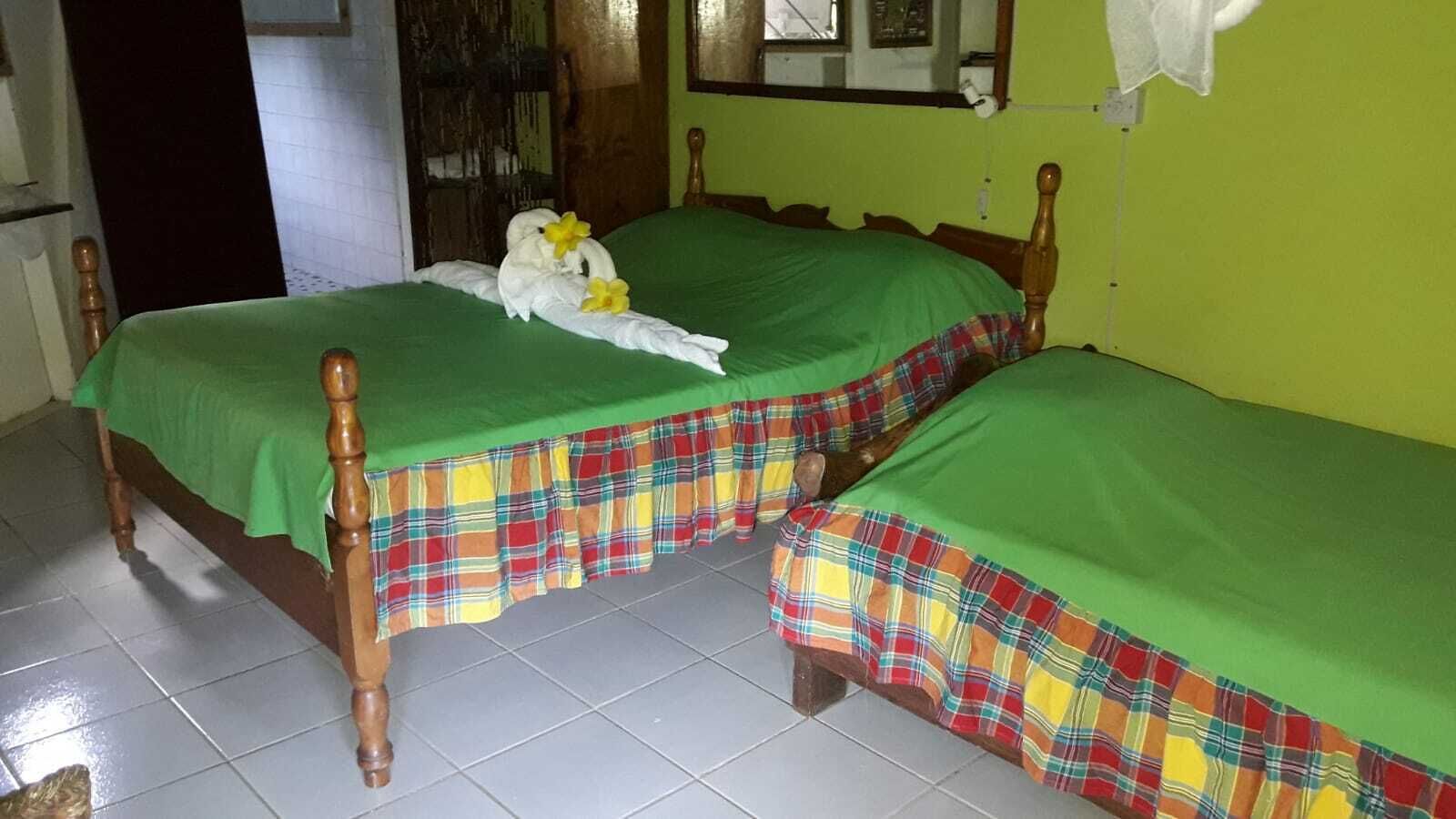 Deux lits simples avec une literie verte et des couvre-lits à carreaux, une tête de lit en bois et un sol carrelé.