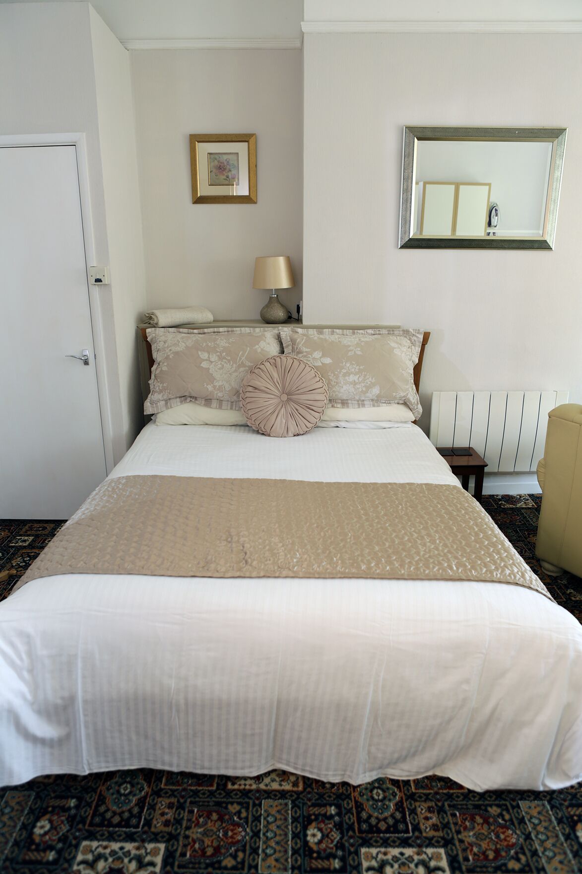 A neatly made bed with a beige bedspread, a floral pillow, and a matching throw blanket. There is a bedside table with a lamp and a framed picture on the wall above the bed.