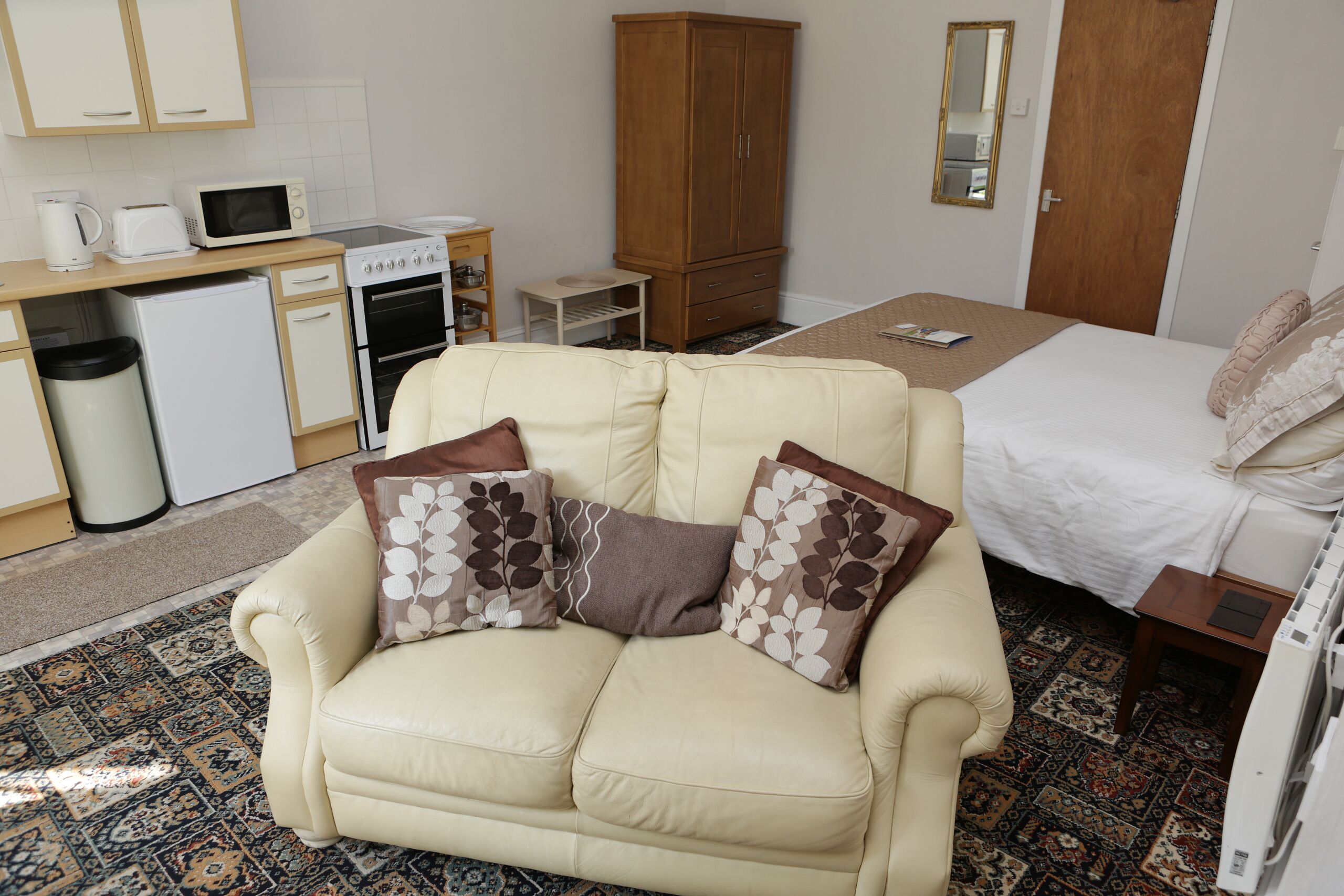 Ein kompaktes Hotelzimmer mit Küchenzeile, einer Couch mit gemusterten Kissen, einem Bett mit weißer Bettdecke und einem Teppich mit Muster.