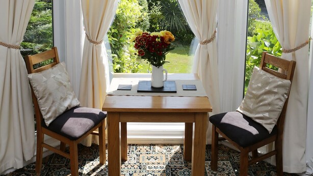 A room with two wooden chairs, a table, and a window with curtains. There is a vase with flowers on the table. - Abbey View Holiday Flats (Torquay)