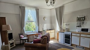 A room with a bed, a desk, a chair, a sofa, a kitchenette, and a window with curtains. - Abbey View Holiday Flats (Torquay)
