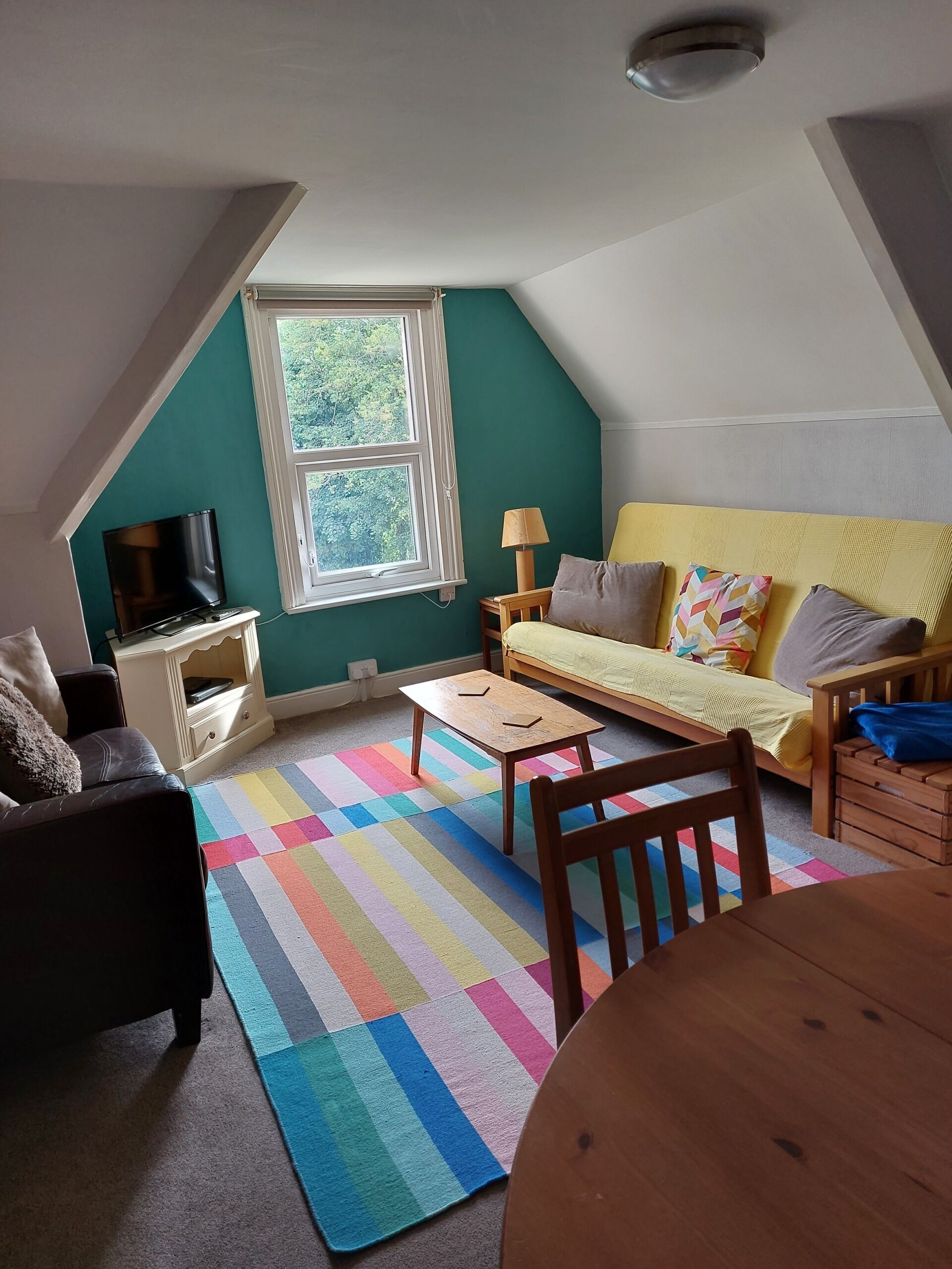 Une pièce confortable de style loft, avec un plafond en pente, une grande fenêtre, un téléviseur, un canapé, une petite table et un tapis coloré.