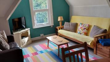 Una habitación acogedora estilo loft, con techo inclinado, un ventanal grande, un televisor, un sofá, una mesita y una alfombra colorida.
