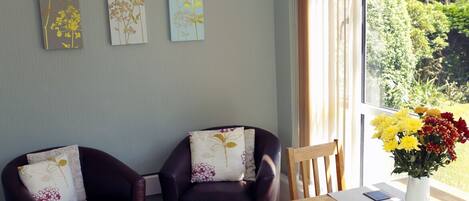 A living room with two leather armchairs, a wooden dining table, and a large window with a view of greenery.