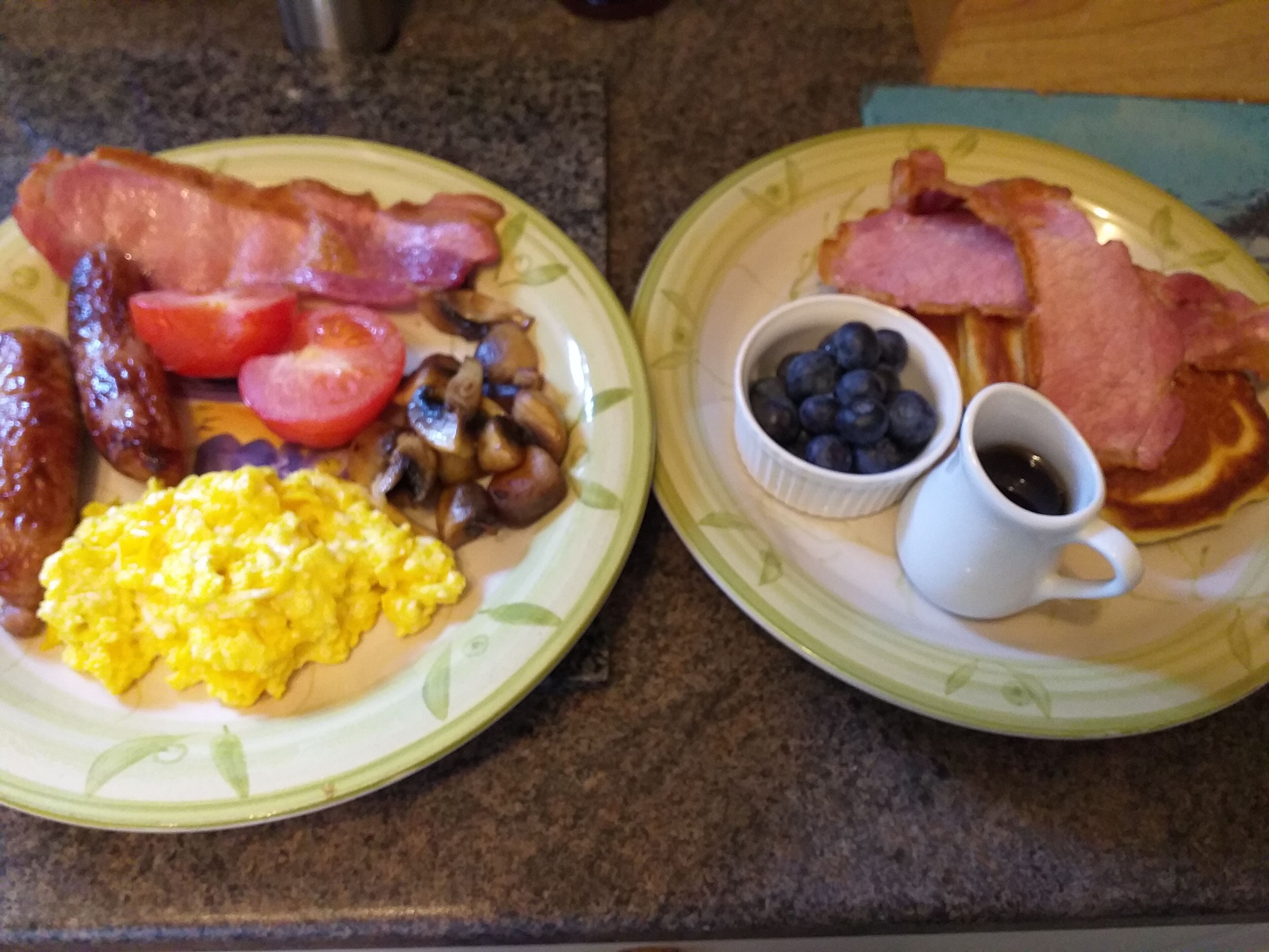 Two plates of breakfast food including bacon, sausages, scrambled eggs, tomatoes, mushrooms, blueberries, and a cup of coffee.