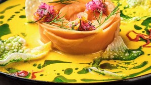 A dish with a yellow sauce, green garnish, and pink flowers on a black plate.