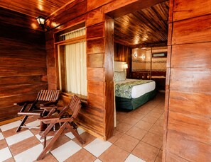A wooden cabin interior with a checkered floor, a bed with a green bedspread, a wooden chair, and a window with curtains. - Hotel Heliconia - Monteverde (Monteverde)