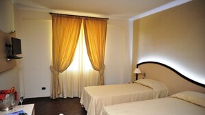 Quarto de hotel com duas camas, televisão, escrivaninha com abajur e janela com cortinas.