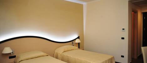 A hotel room with two single beds, a nightstand with a lamp, and a door leading to another room.