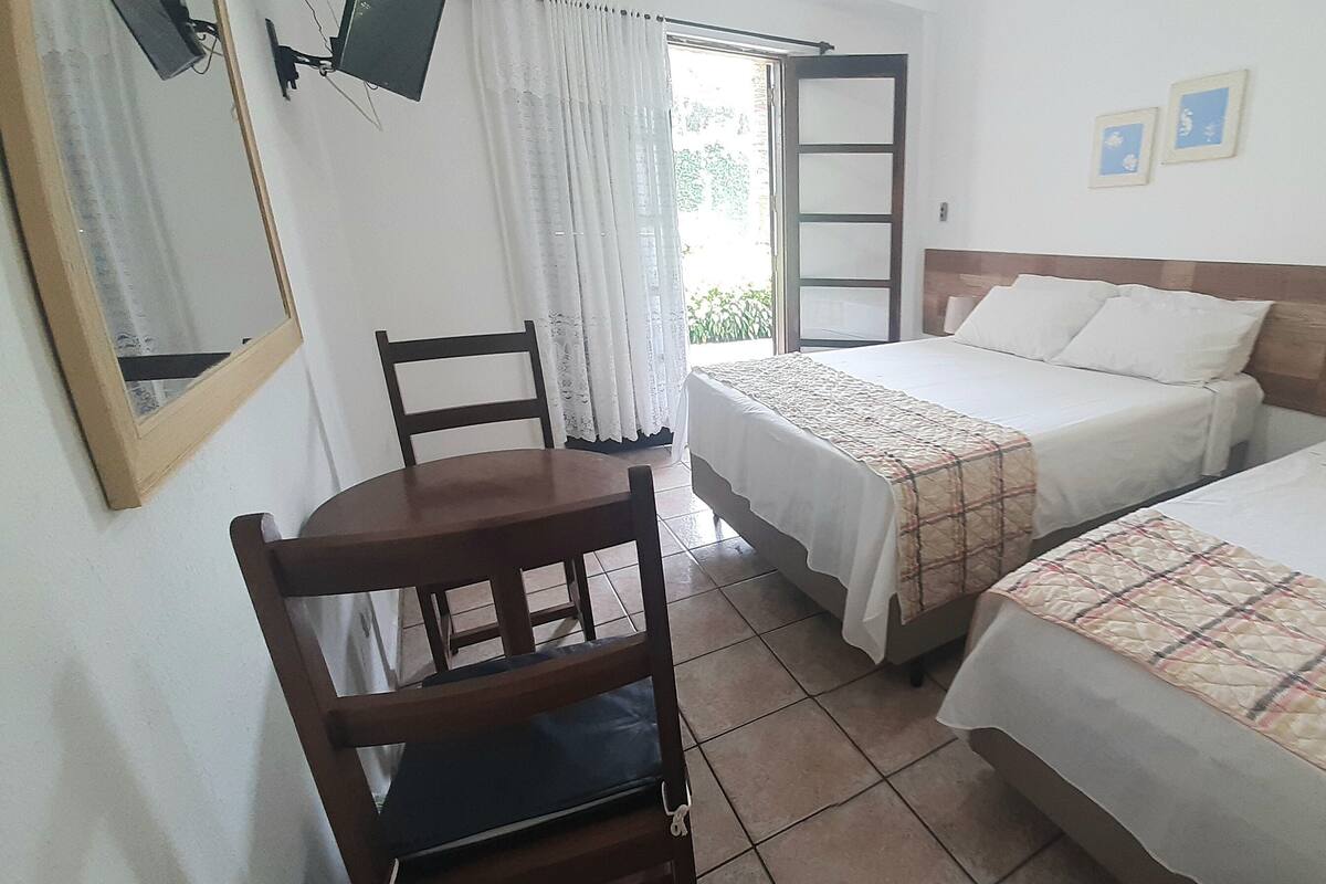 Habitación de hotel con dos camas, una silla de madera, una mesita y un ventanal con cortinas.