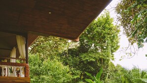 A covered outdoor seating area with a white sofa, a wooden table with a plant, and a person holding a striped bag. - Hotel Villabosque Eco Boutique (Quepos)