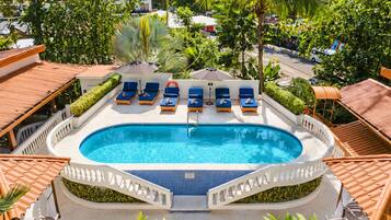 Una piscina con sillas de descanso y sombrillas, rodeada de vegetación y edificios.