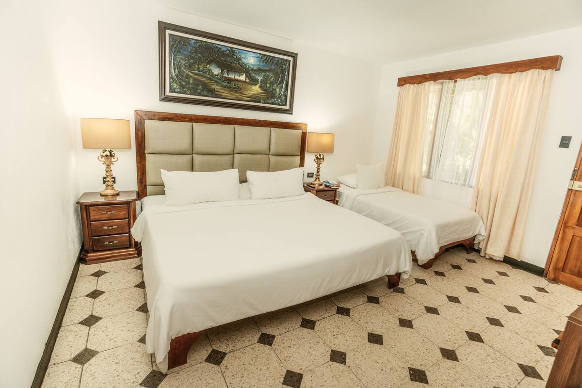 Une chambre d’hôtel avec un grand lit, deux tables de chevet, une commode, un tableau au mur et une fenêtre avec des rideaux.
