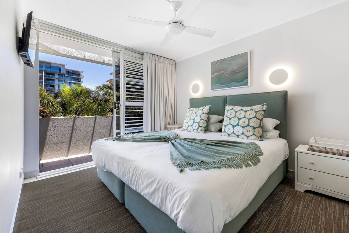 A modern bedroom with a large bed, a ceiling fan, a nightstand, and a view of the outside balcony.