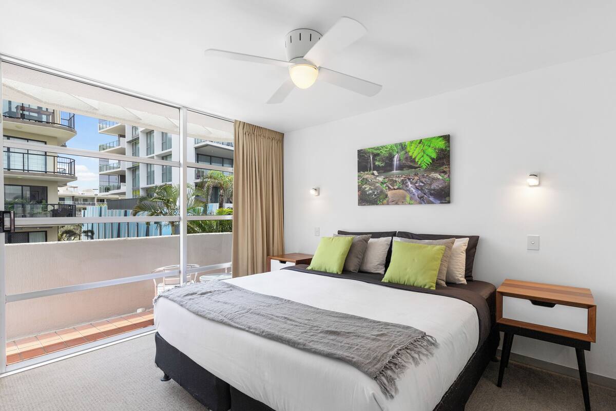 A modern hotel room with a large bed, a balcony with a view of the building, a ceiling fan, and a painting on the wall.