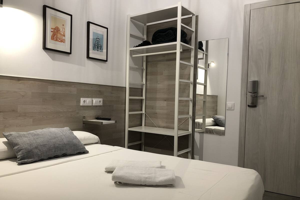 A modern hotel room with a bed, a white wardrobe, a built-in shelf unit, and two framed pictures on the wall.