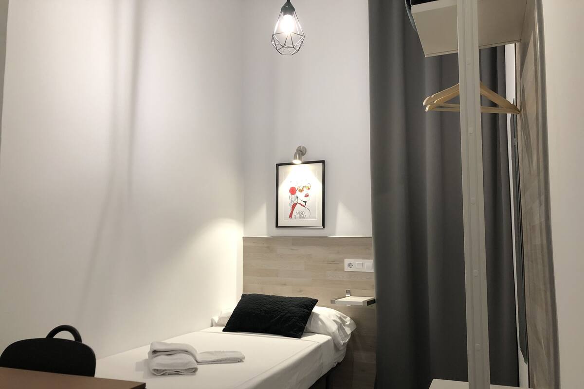 A hotel room with a single bed, a desk, a chair, a hanging light fixture, and a framed picture on the wall.