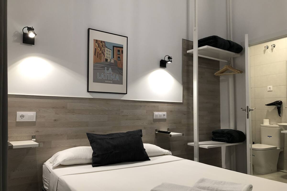 A modern hotel room with a bed, a nightstand, a framed picture on the wall, a wardrobe, and a bathroom with a toilet and sink.
