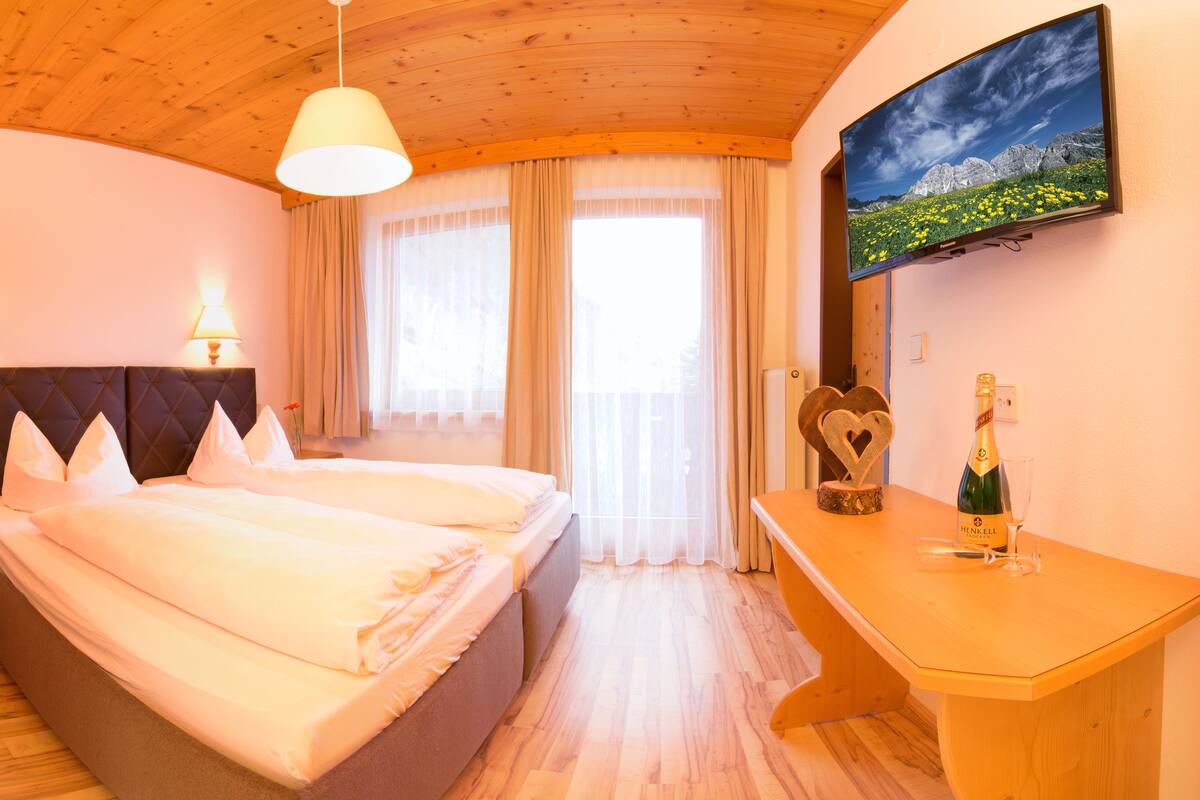 A hotel room with a bed, bedside lamps, a TV, a table with a bottle and glass, and a window with curtains.