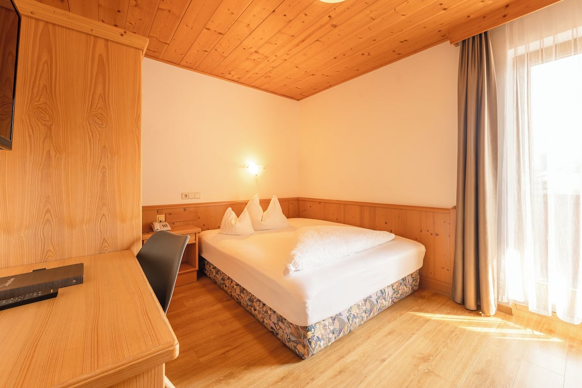 A hotel room with a wooden ceiling, a bed with white bedding, a desk with a remote control, and a window with curtains.