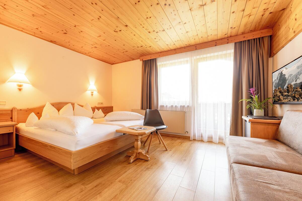 A hotel room with a wooden ceiling, a bed with white bedding, a wooden nightstand, a wooden table, a black chair, a beige sofa, and a window with curtains.