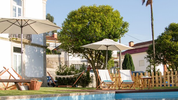 Un área de piscina con sillones de descanso y sombrillas, una palmera y un edificio con techo de tejas.
