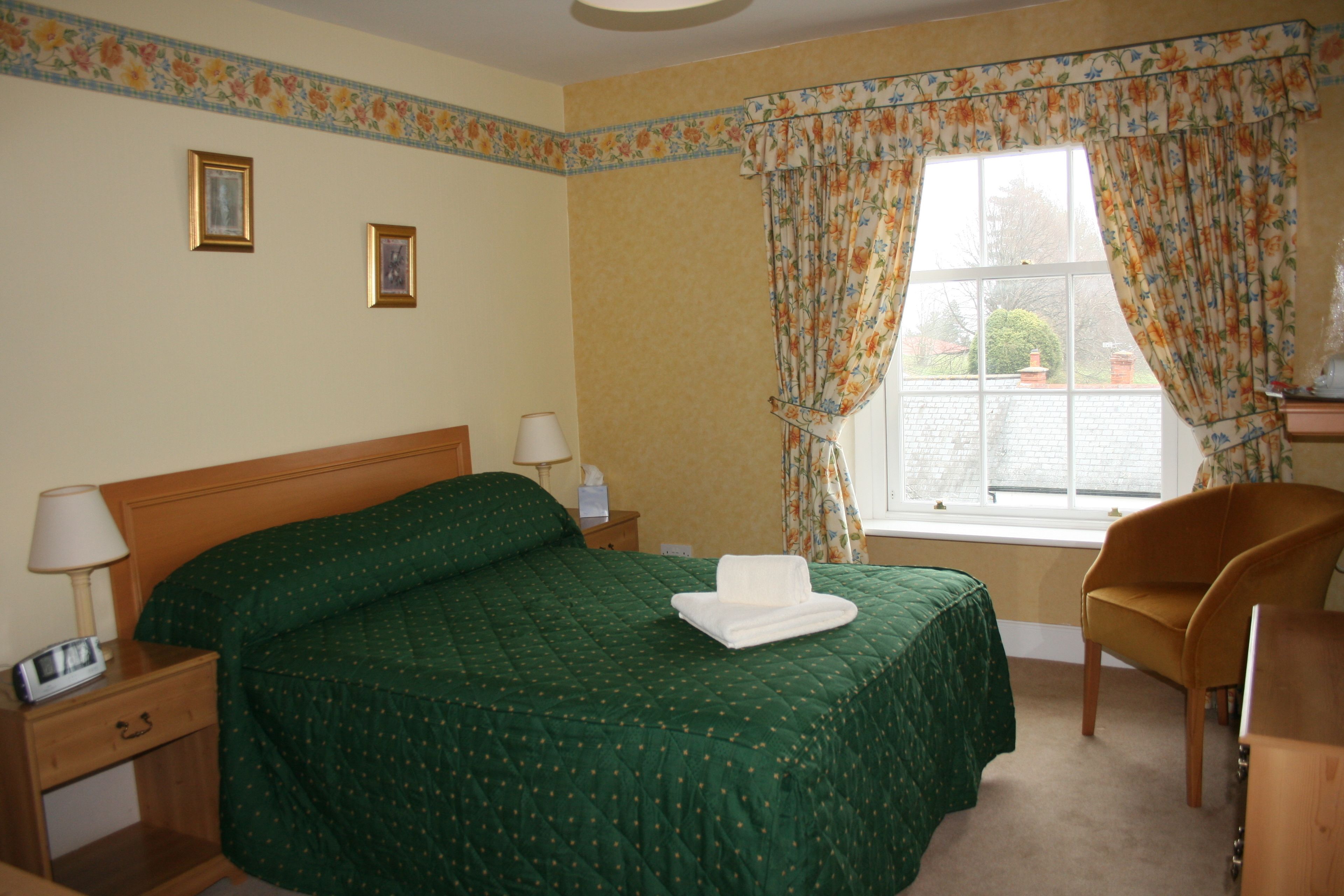 A hotel room with a large bed, a wooden nightstand, a chair, a window with curtains, and a small table.