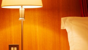 Quarto de hotel com parede de madeira, luminária de cabeceira, cama com lençóis brancos e mesa de cabeceira com interruptor.