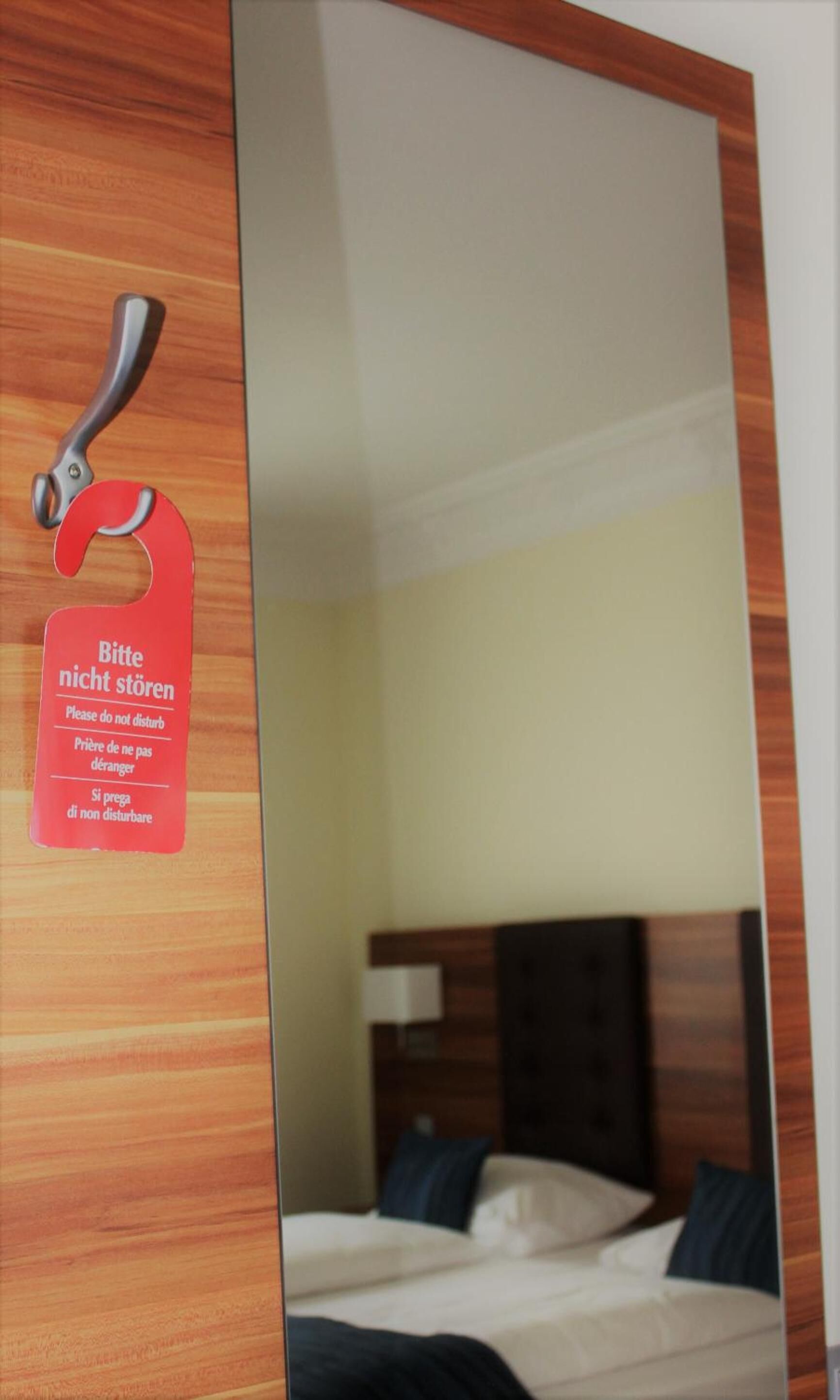 Habitación de hotel con puerta de madera, un letrero rojo con indicaciones en varios idiomas y una cama con sábanas blancas y almohadas oscuras.