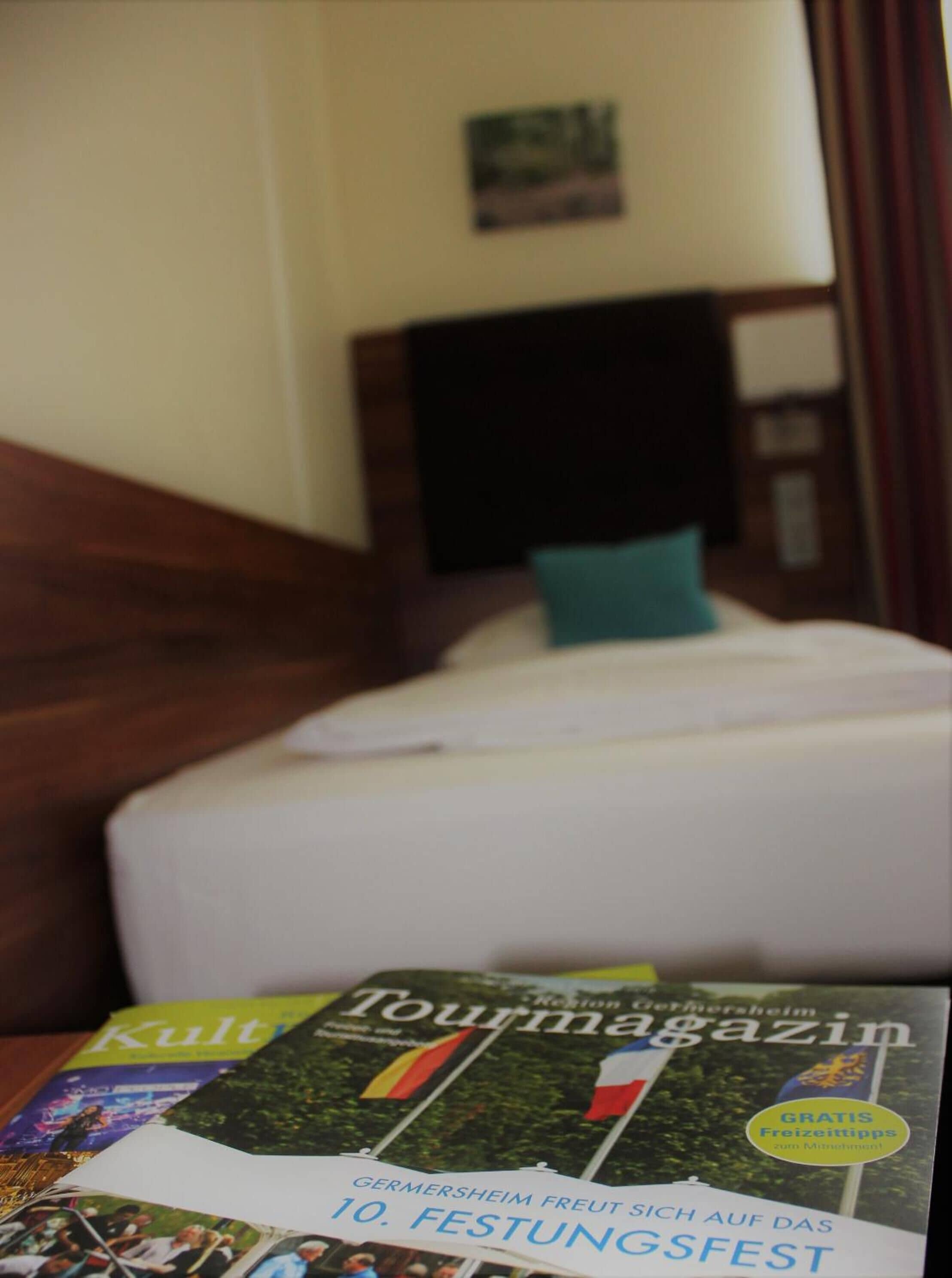 A hotel room with a bed, a wooden headboard, and a pillow. There is a magazine on the bed with the title "Tourmagazin".