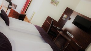 Quarto de hotel com cama, escrivaninha, cadeira, televisão e um quadro na parede.