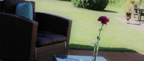 A wicker chair with a cushion and a vase with a single flower on a table.