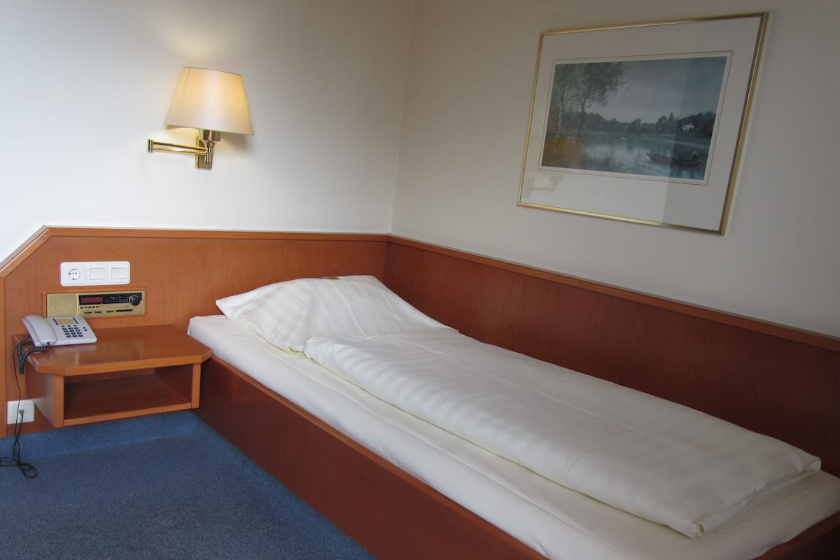 Ein Einzelbett mit Holz-Kopfteil, ein Nachttisch mit Lampe, ein Telefon und ein gerahmtes Bild an der Wand.