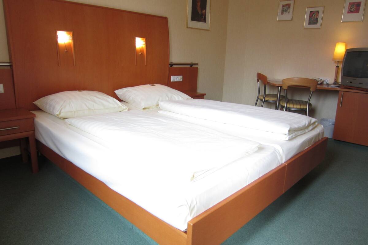 Ein Doppelbett mit zwei Kissen, ein Holzkopfteil, ein Nachttisch, ein Schreibtisch mit Stuhl und ein Fernseher.