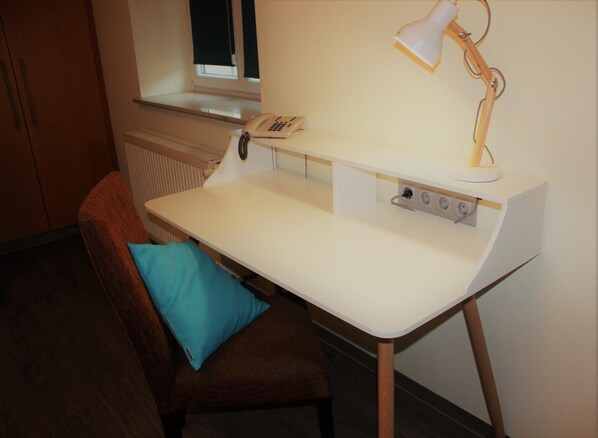 A small, well-lit room with a white desk, a chair with a blue cushion, a lamp, and a telephone on the desk. - Hotel Germersheimer Hof (Germersheim)