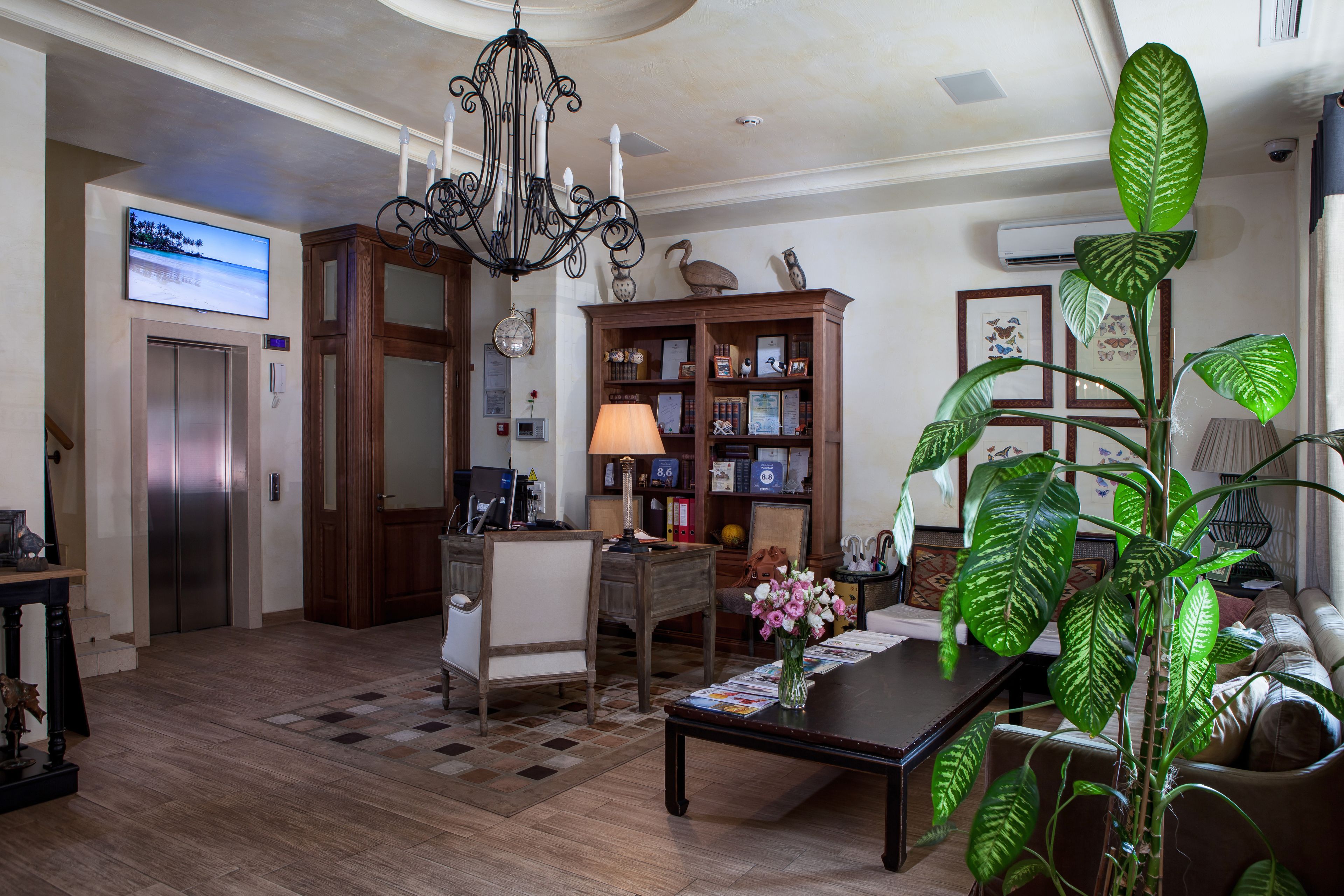 A spacious living room with a chandelier, a large green plant, a wooden cabinet with decorative items, and a television mounted on the wall.
