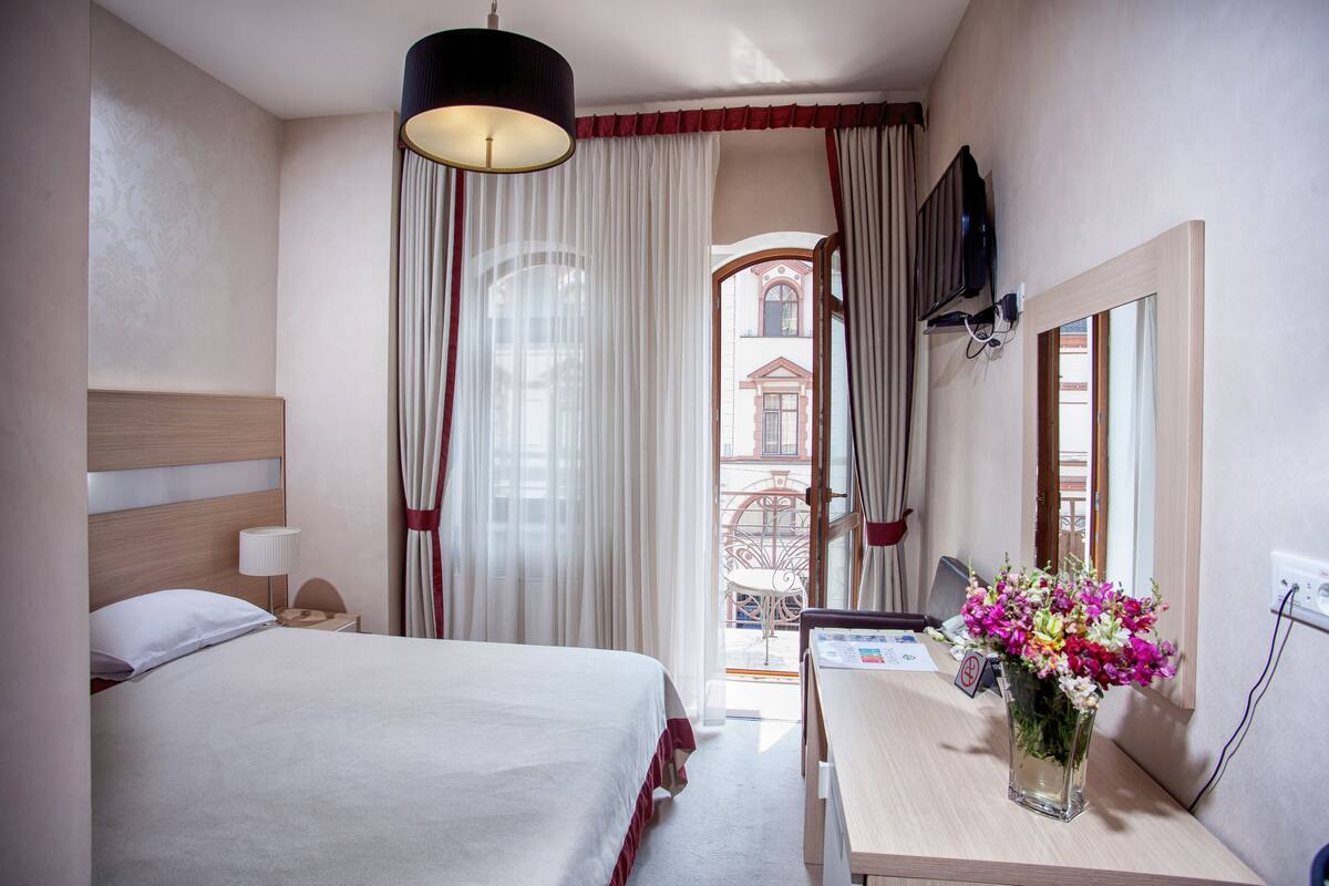 A hotel room with a bed, a desk with a vase of flowers, a television, and a view of the outside.