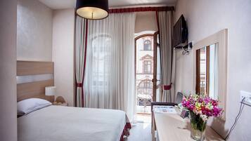 A hotel room with a bed, a desk with a vase of flowers, a television, and a view of the outside.