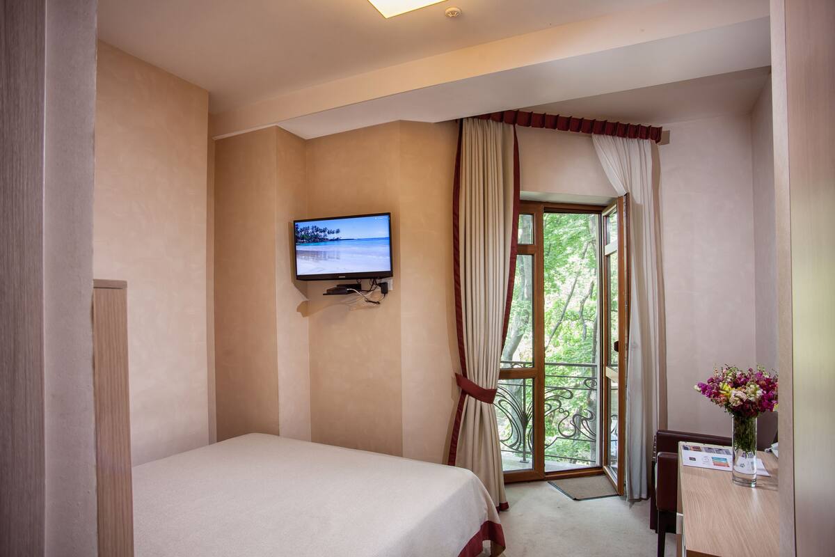 A hotel room with a bed, a TV mounted on the wall, a window with curtains, and a table with a vase of flowers.