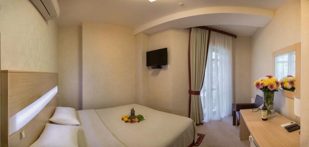 A hotel room with a bed, a TV, a desk with a vase of flowers, and a window with curtains.