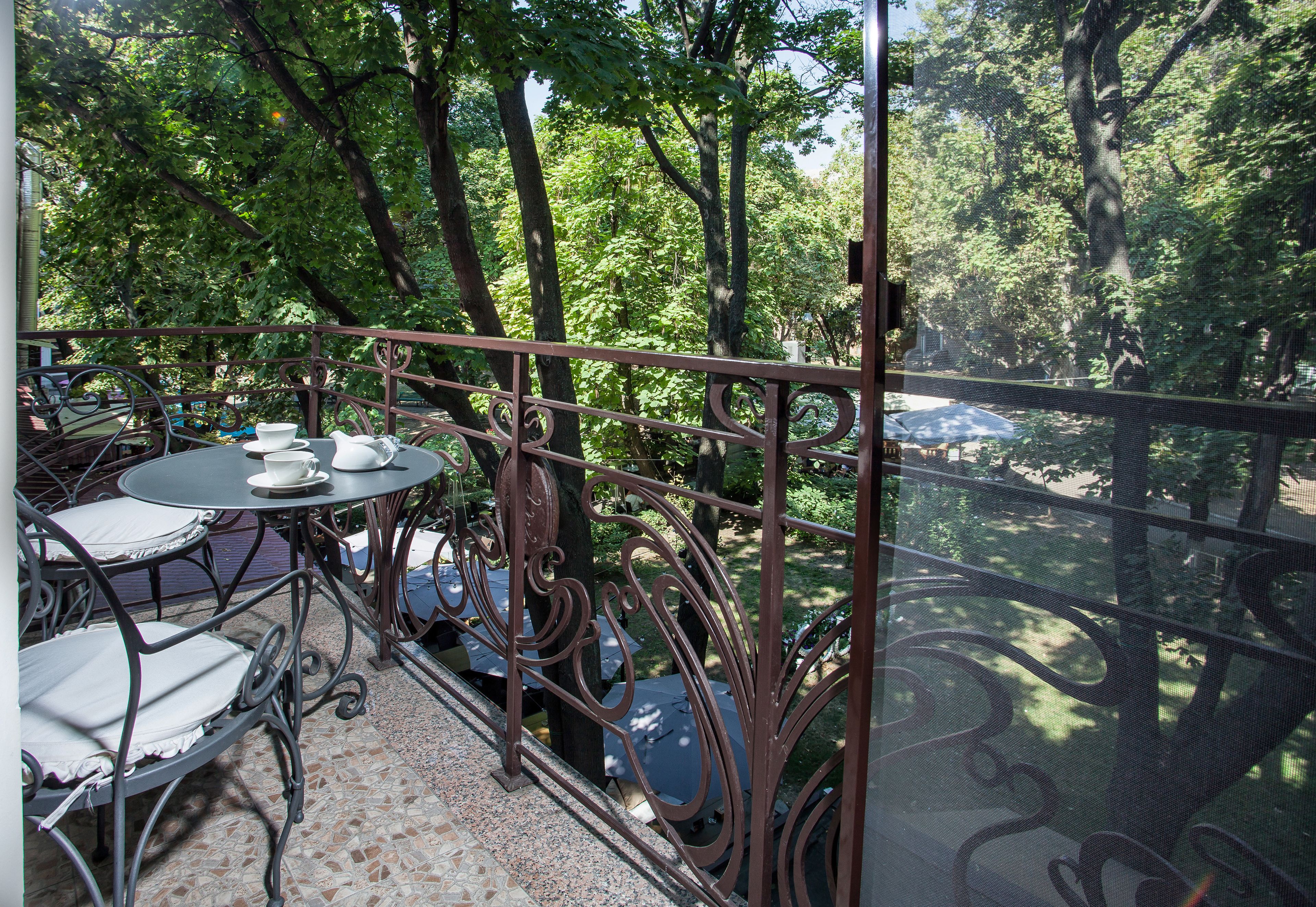 A balcony with a wrought iron railing, a round table with chairs, and a view of a lush green park.